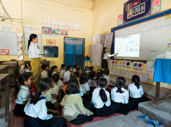 Young children in a classroom with their teacher, and a girl pointing out something projected onto the board.